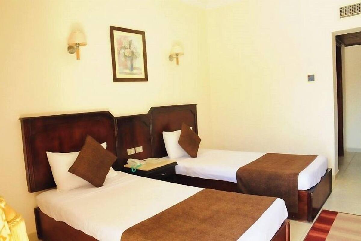 A hotel room with two beds, each with a wooden headboard, white linens, and brown throw blankets. There are two wall-mounted lamps and a framed picture on the wall.