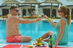 Deux personnes assises au bord d’une piscine, avec un verre et une bouteille d’alcool.