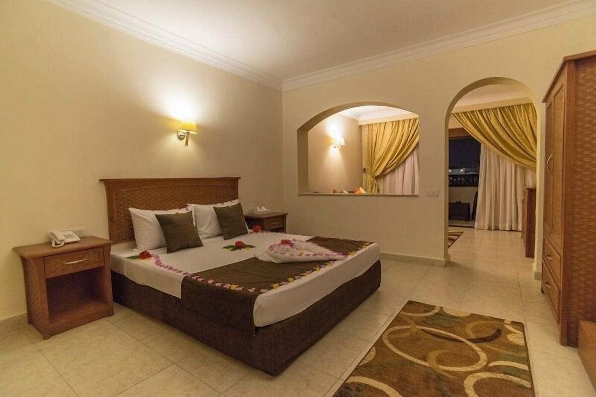 A hotel room with a large bed, a nightstand, a bedside lamp, a mirror, and a view of the outside.