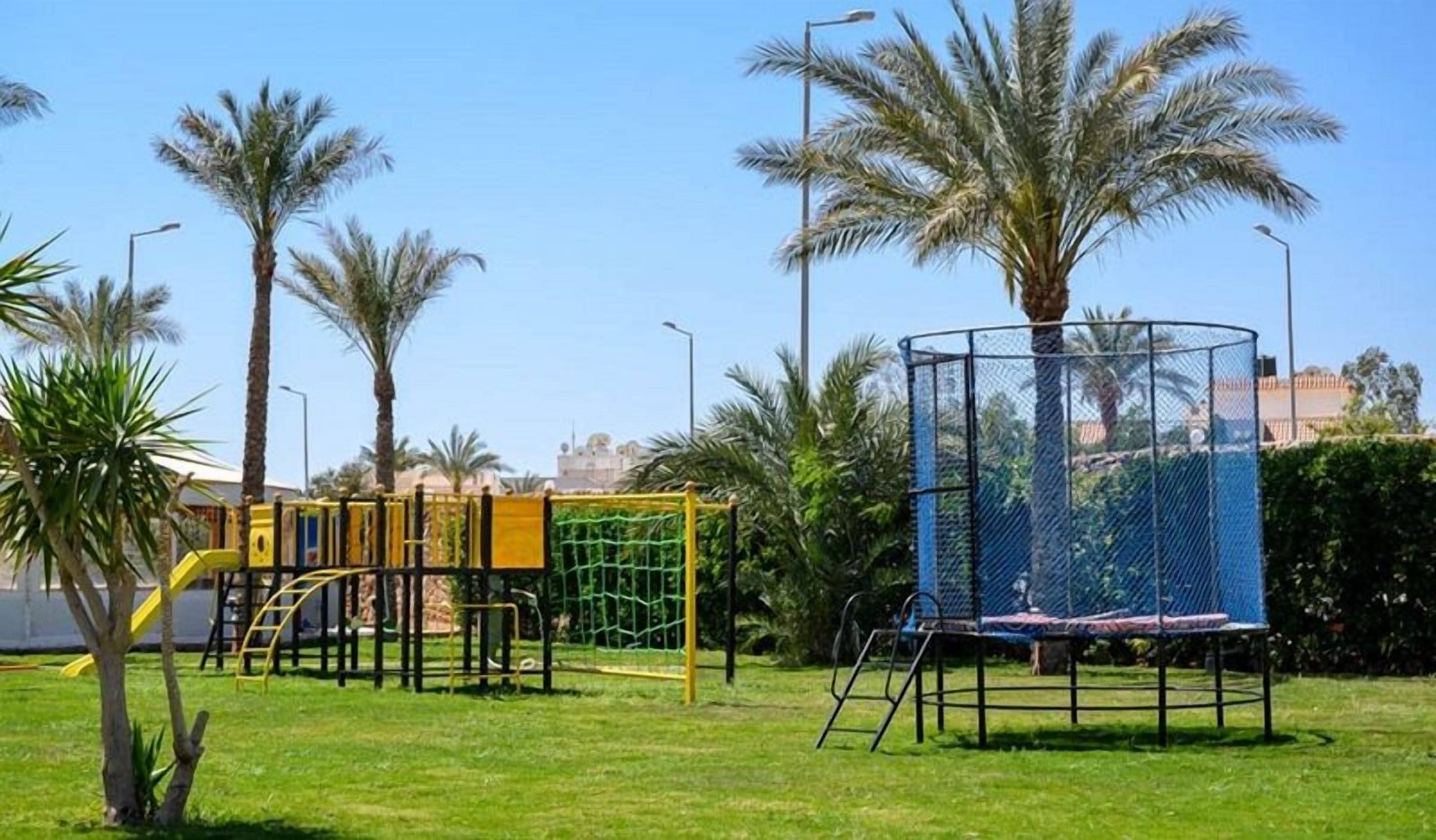 A playground with various play structures, including a climbing frame and a swing set, surrounded by palm trees and a clear sky.