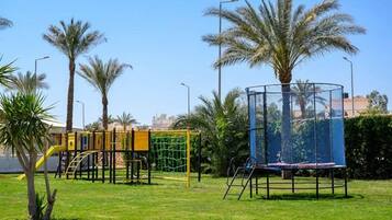 A playground with various play structures, including a climbing frame and a swing set, surrounded by palm trees and a clear sky.