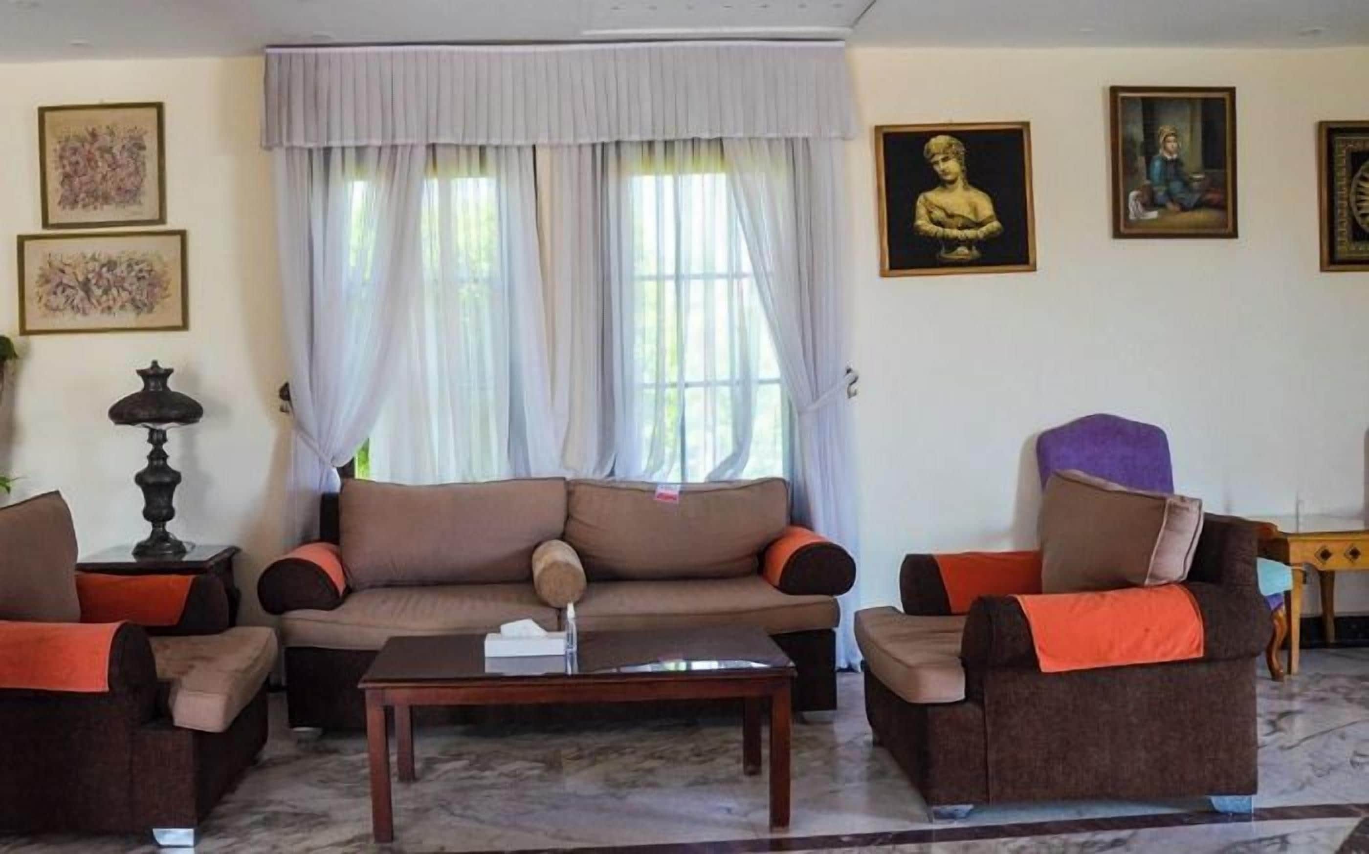 A living room with a sofa, two armchairs, a coffee table, and a lamp. There are two windows with curtains, a wall with framed artwork, and a statue.