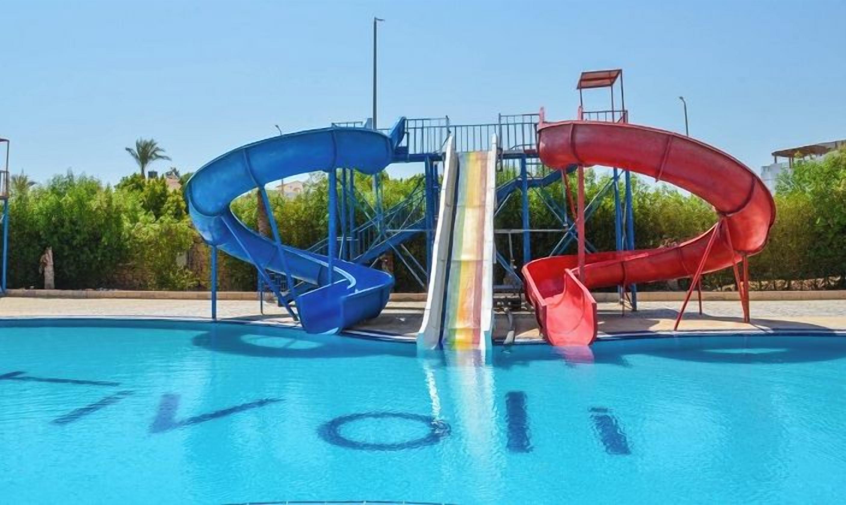 A swimming pool with three slides, a clear sky, and greenery in the background.