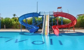 A swimming pool with three slides, a clear sky, and greenery in the background.
