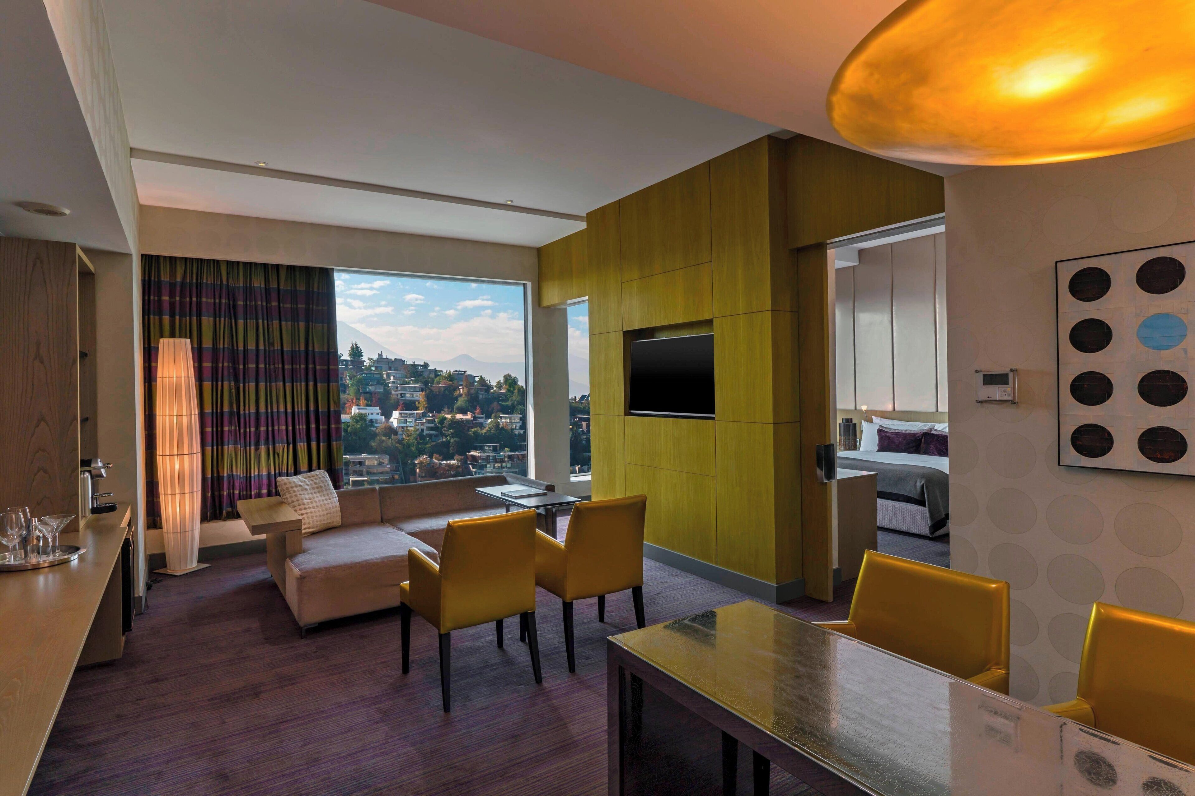 Une chambre d’hôtel moderne équipée d’un canapé, d’une table à manger, de chaises, d’un téléviseur et d’une grande fenêtre donnant sur la ville.