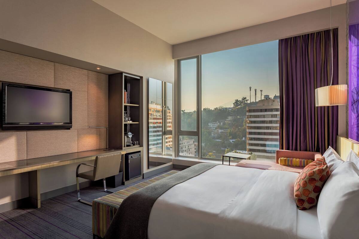 A modern hotel room with a large bed, a desk, a TV, and a view of the cityscape.