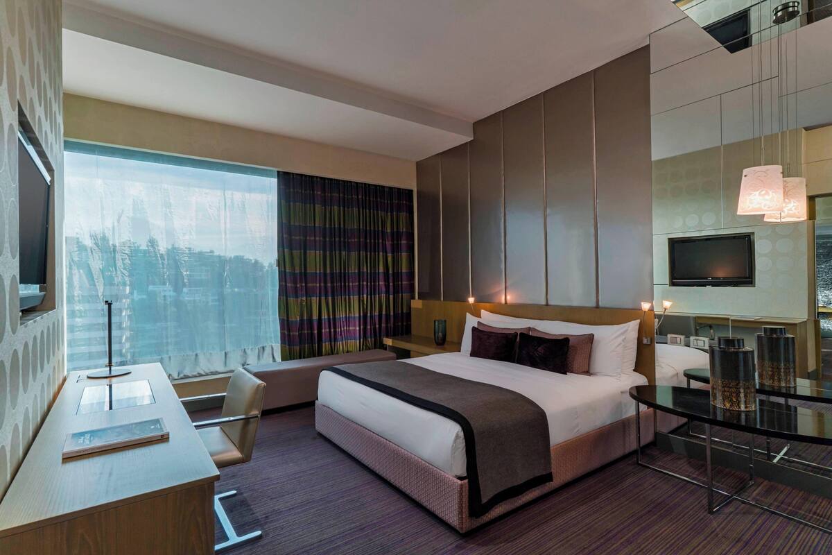 A modern hotel room with a large bed, a desk, a chair, a TV, and a window with curtains.