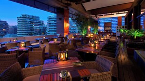 Un restaurante en la azotea con mobiliario de mimbre, mesas con velas y vistas a edificios de la ciudad.