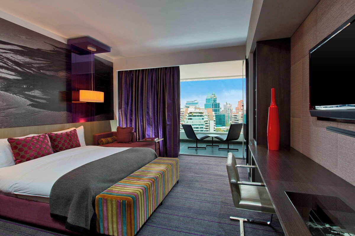 A modern hotel room with a large bed, a colorful bench, a desk, and a city view.