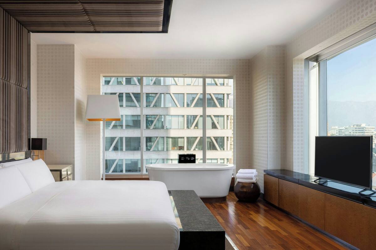 A modern hotel room with a large bed, a bathtub, a television, and a view of a cityscape through large windows.