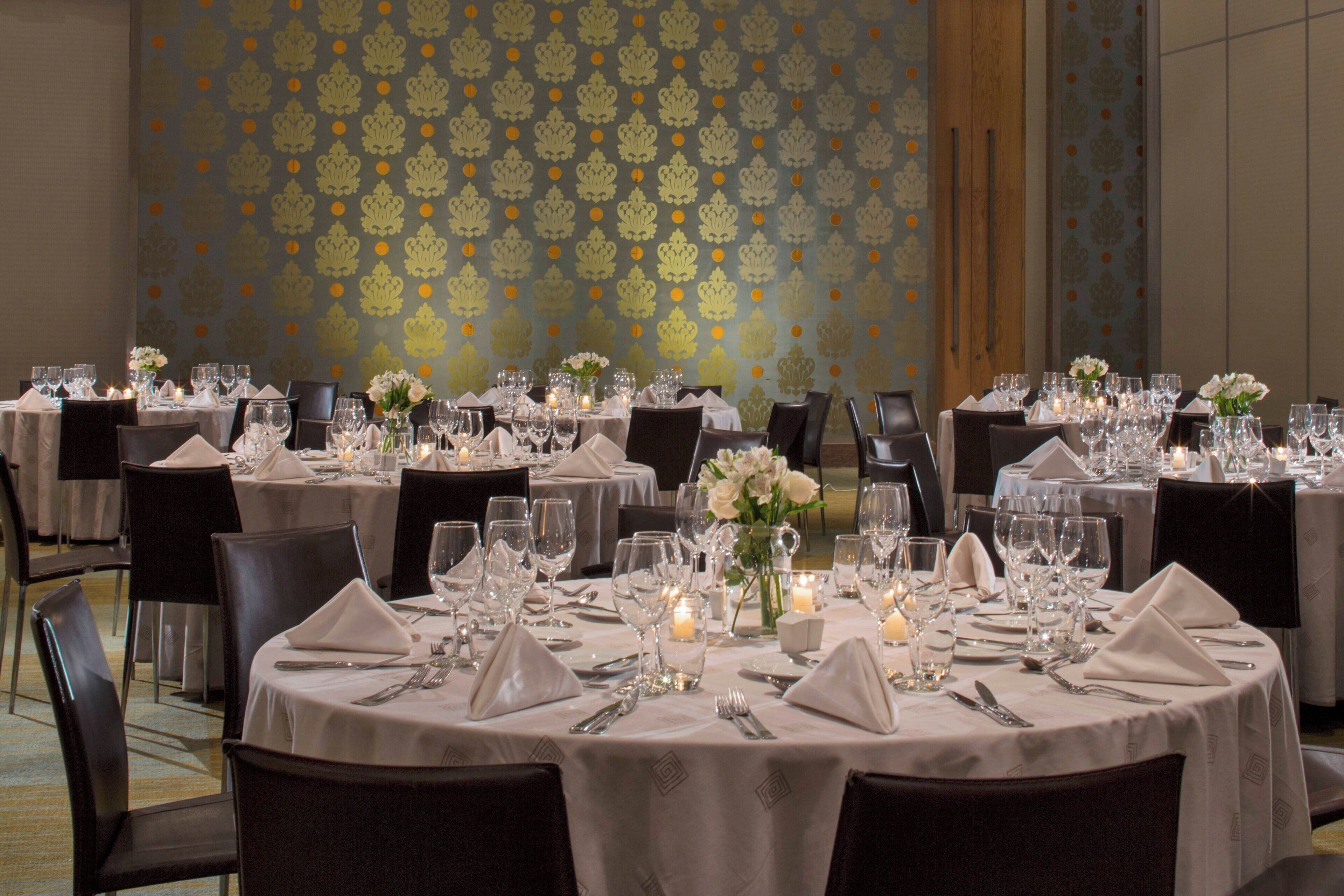 A banquet hall with round tables set for dining, each with white tablecloths, candles, and floral centerpieces.