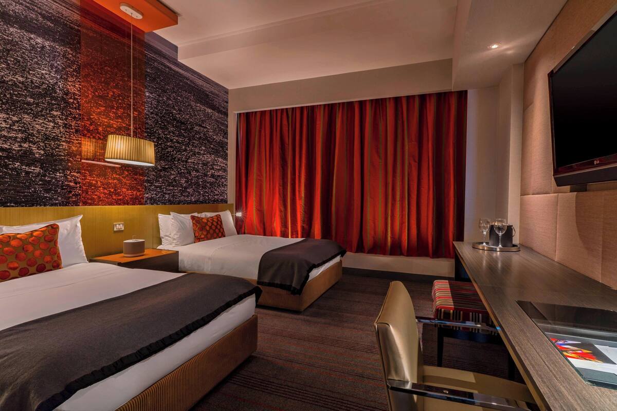 A hotel room with two beds, a desk, a TV, and a large window with red curtains.