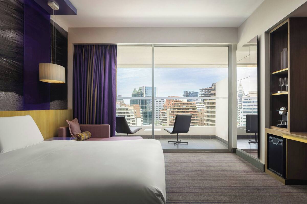A hotel room with a large bed, two chairs, a desk, and a view of the cityscape through large windows.
