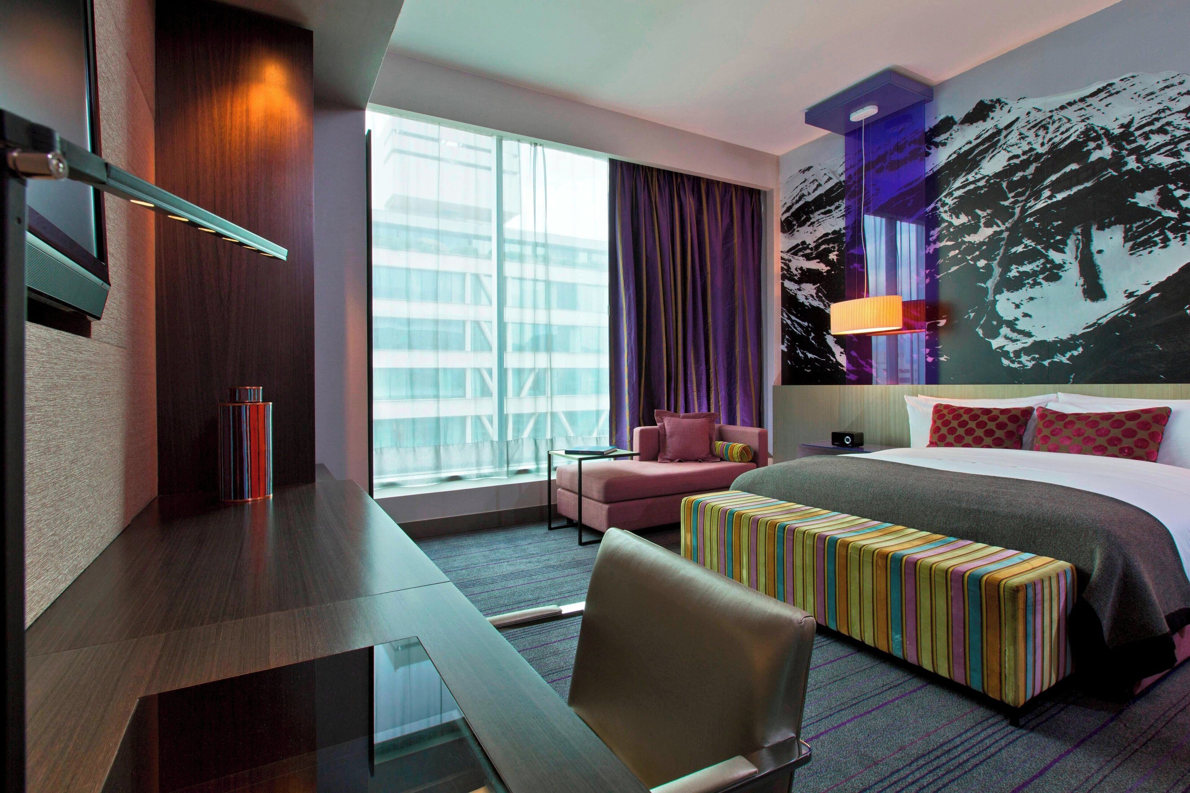 A modern hotel room with a large bed, a desk, a chair, a small table, a colorful bench, a window with curtains, and a wall-mounted lamp.