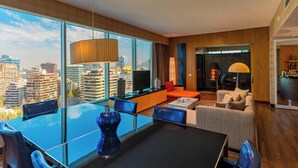Una habitación de hotel moderna con una amplia vista de la ciudad, un comedor con mesa de vidrio y una zona de estar acogedora con sofá y sillones.