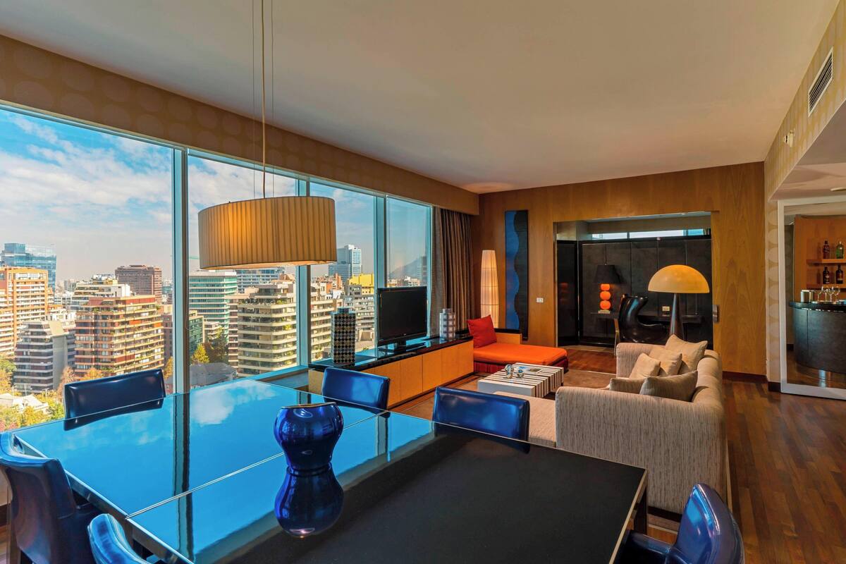 A modern hotel room with a large city view, a dining area with a glass table, and a cozy seating area with a sofa and armchairs.