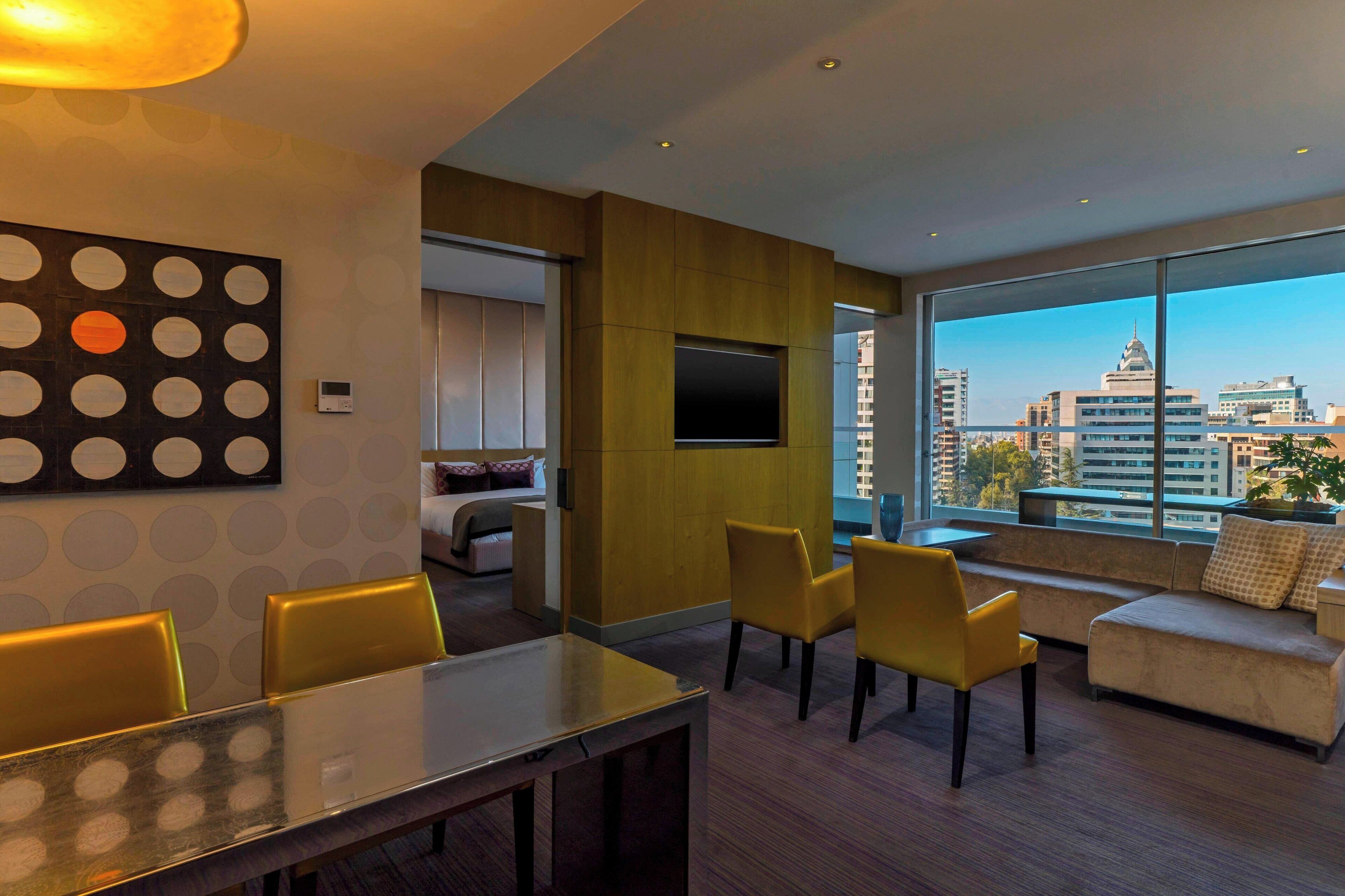 A modern hotel room with a dining area, a sofa set, and a large window offering a city view.
