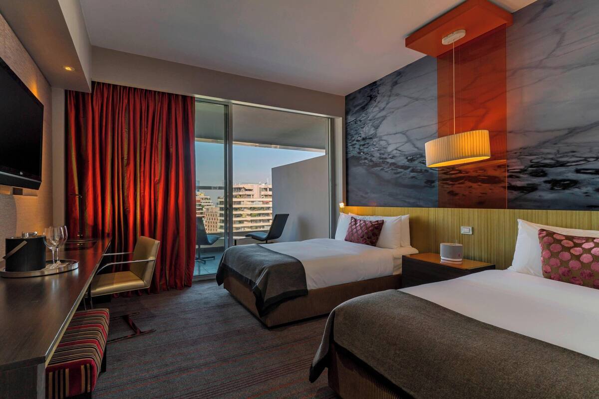 A hotel room with two beds, a desk with a chair, a TV, a large window with curtains, and a city view.