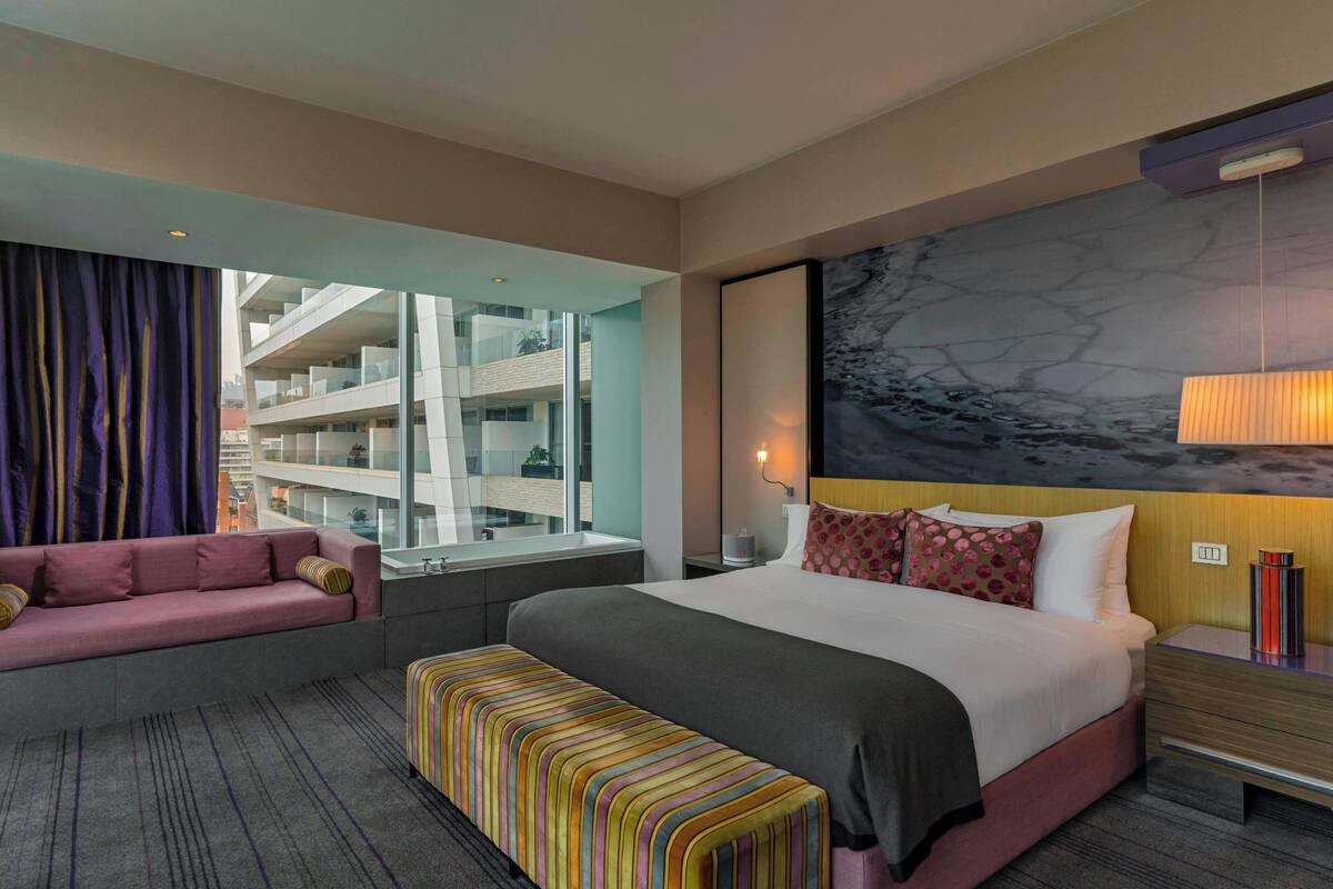 A hotel room with a bed, a sofa, a bench, a large window, and a view of a building.