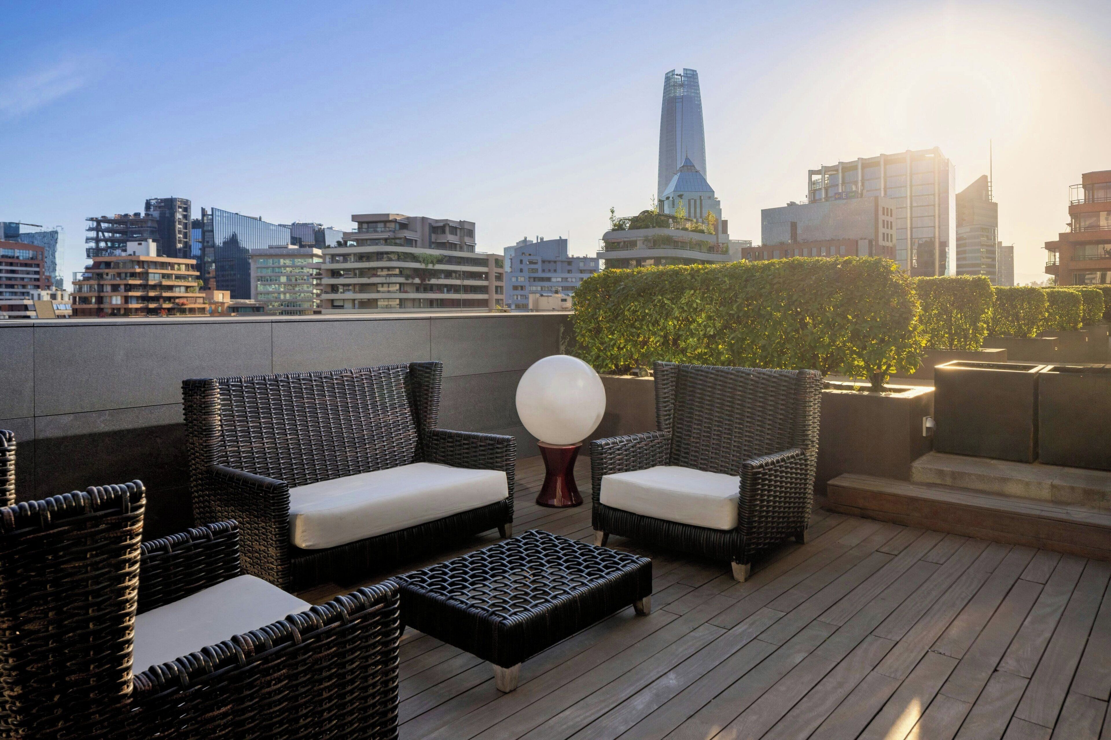 A rooftop terrace with wicker furniture and a spherical decorative object, overlooking a cityscape with modern buildings.