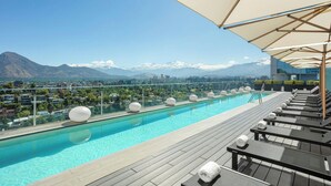 Una piscina en la azotea con sillones de descanso y sombrillas, que ofrece vistas a un paisaje urbano y montañas.