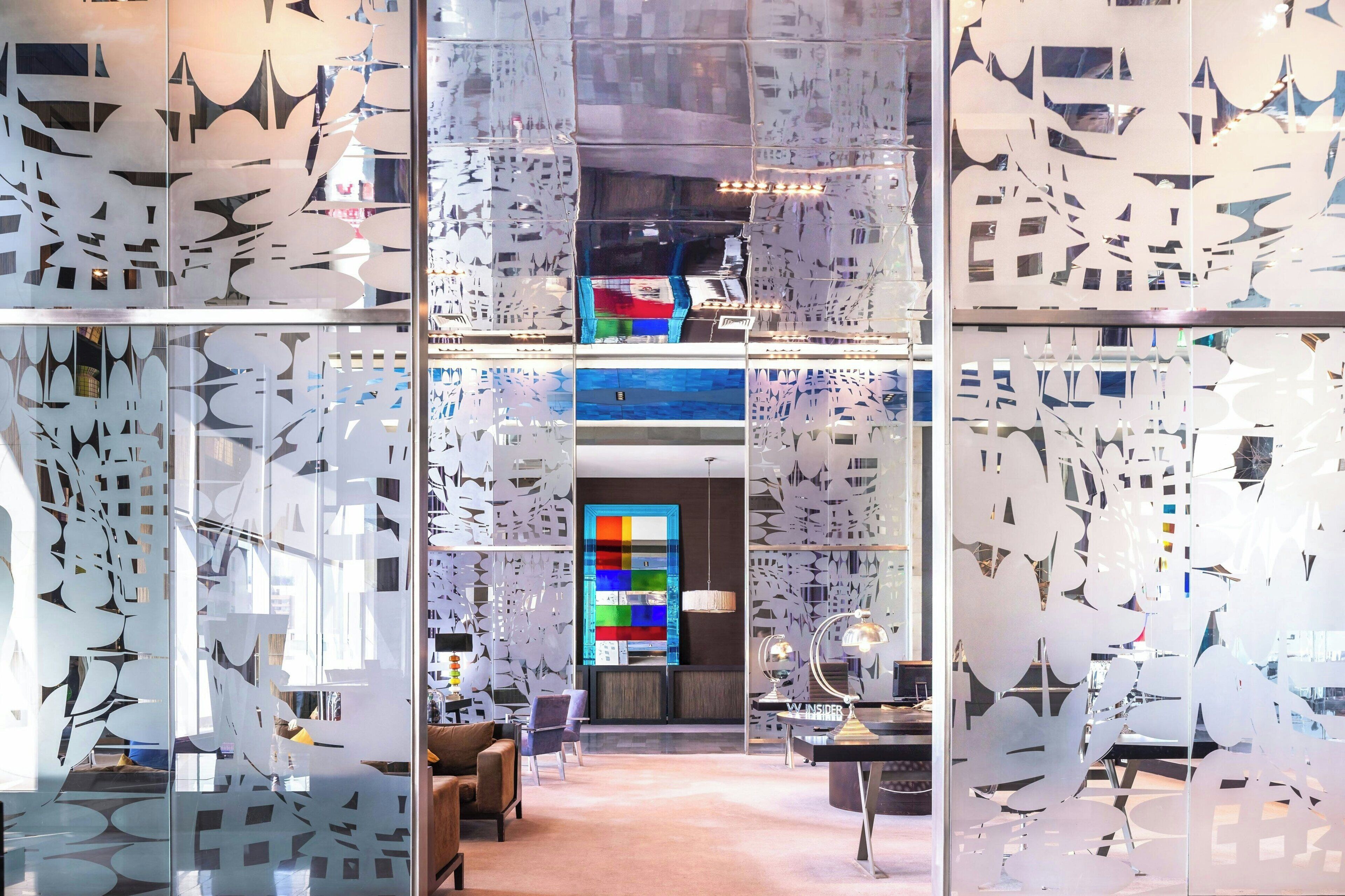 A modern hotel lobby with a glass partition featuring abstract cut-out designs, a colorful abstract art piece on the wall, and a reception desk with a lamp.