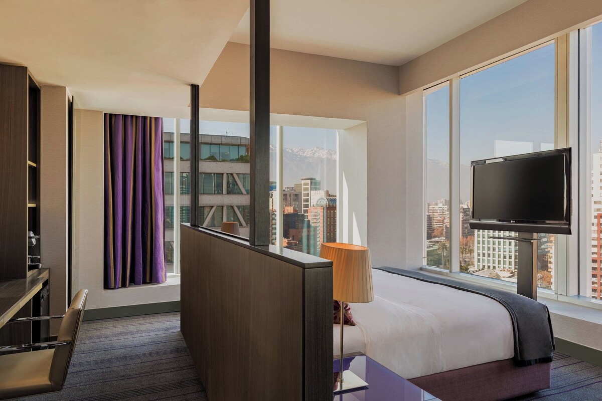 A hotel room with a large window, a bed, a TV, and a desk.