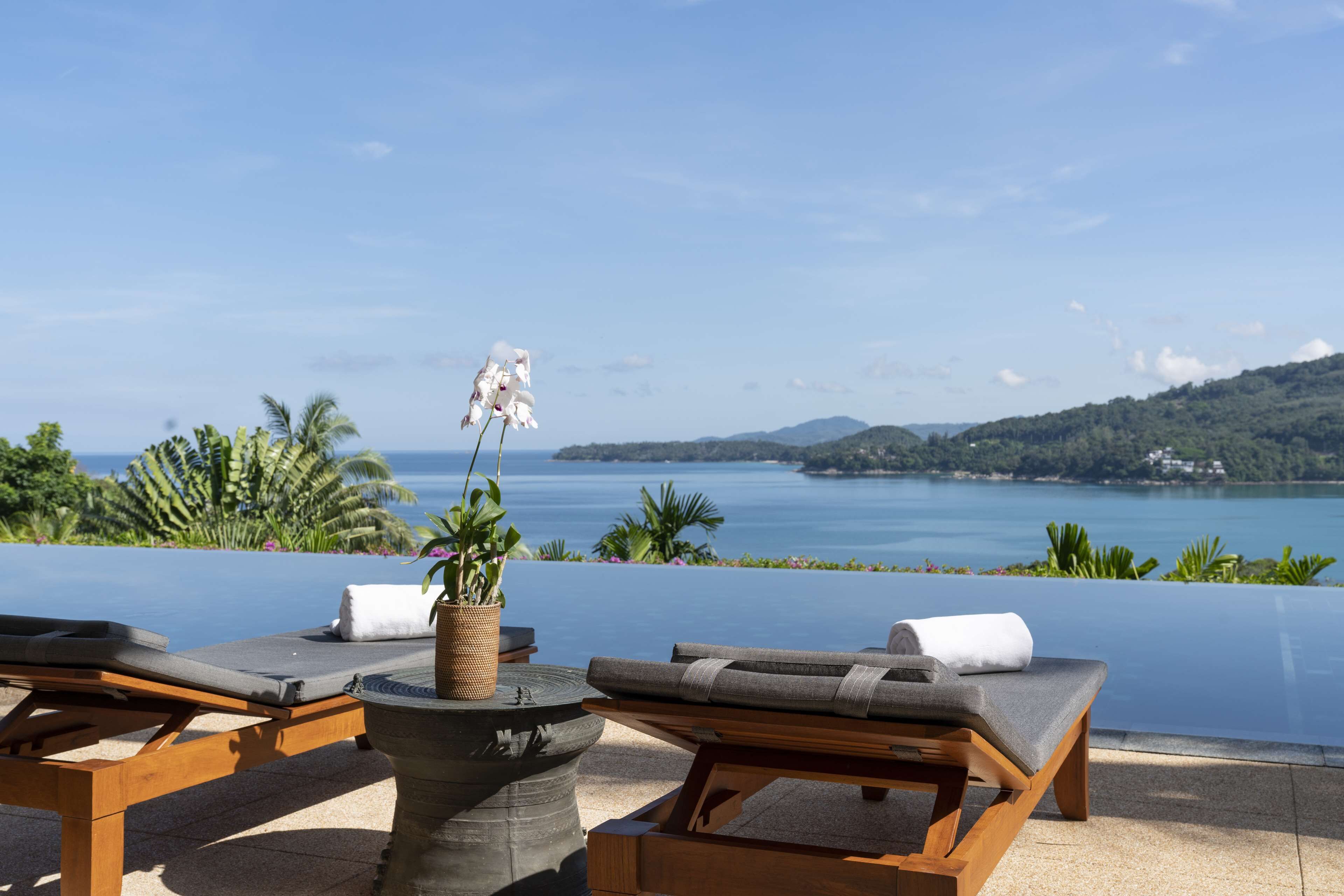 A poolside lounge area with two wooden lounge chairs, a stone table with a woven basket, and a potted orchid.