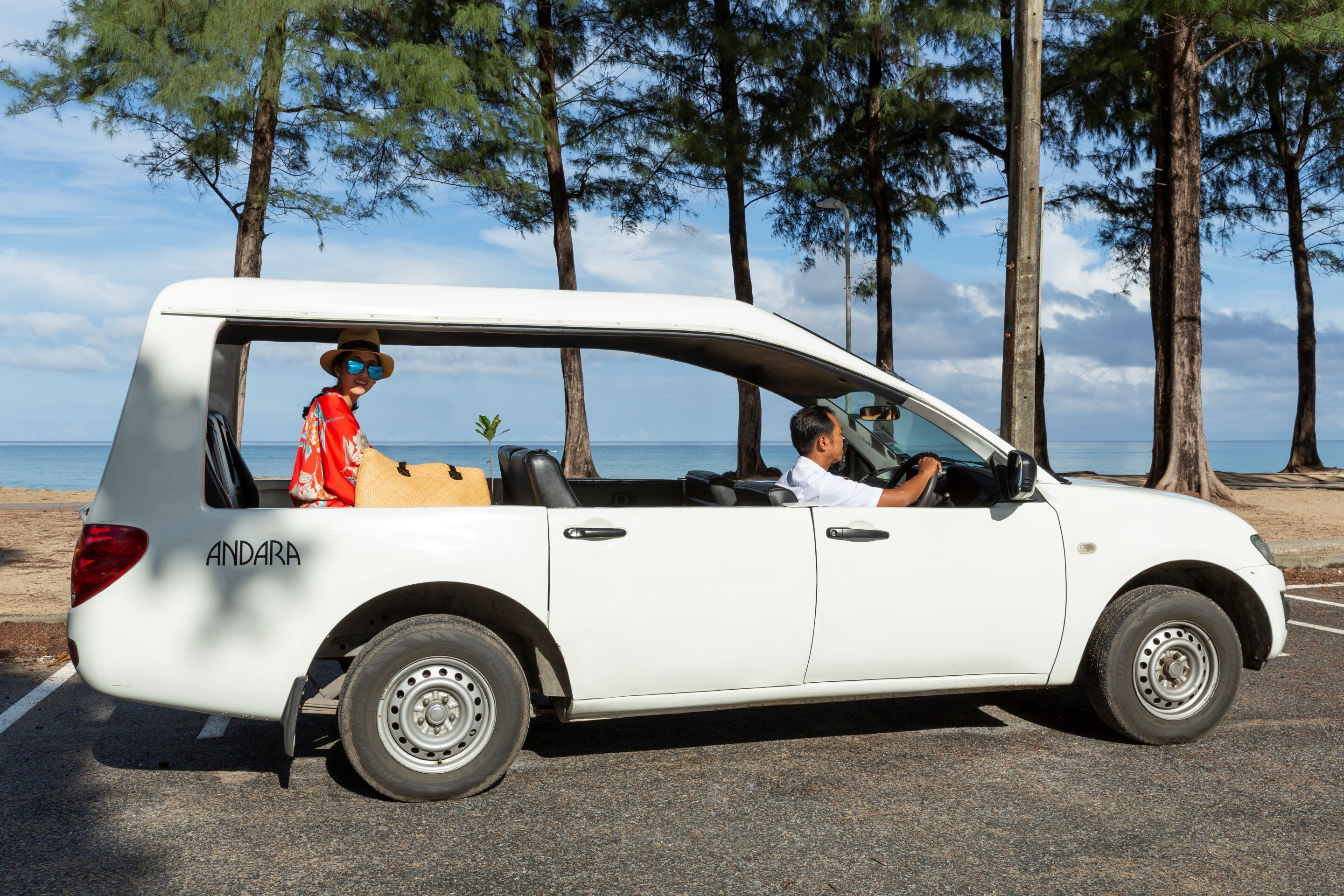 A white convertible car with the name "ANDARA" on the back, parked on a road with trees and a body of water in the background.