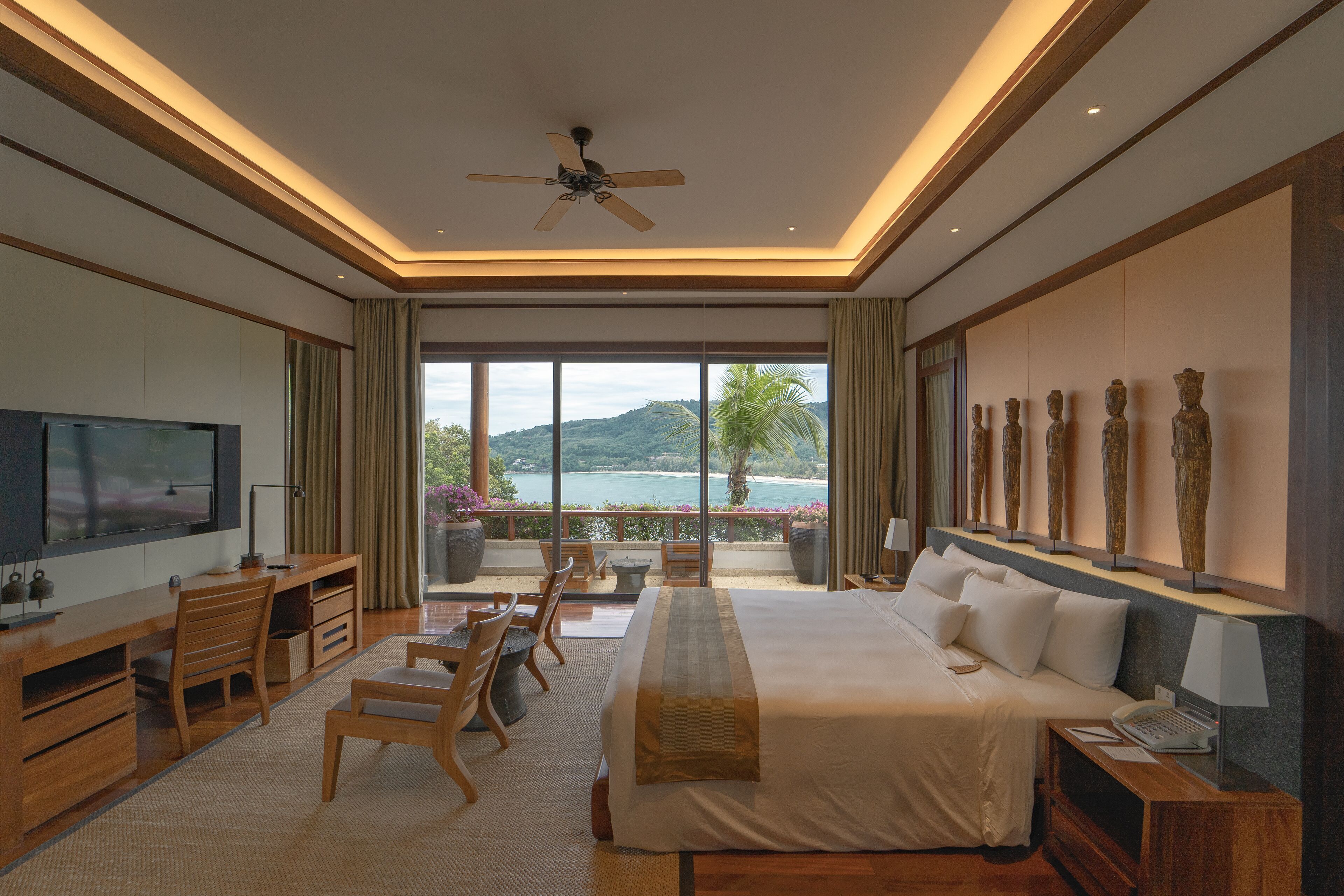 A modern hotel room with a large bed, a desk, a television, and a balcony with a view of the sea.