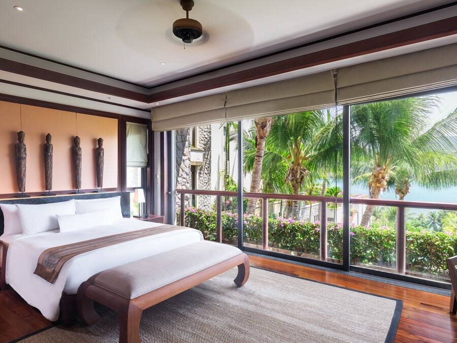 Un dormitorio amplio con una cama grande, un banco de madera y un balcón con vistas a palmeras y una masa de agua.