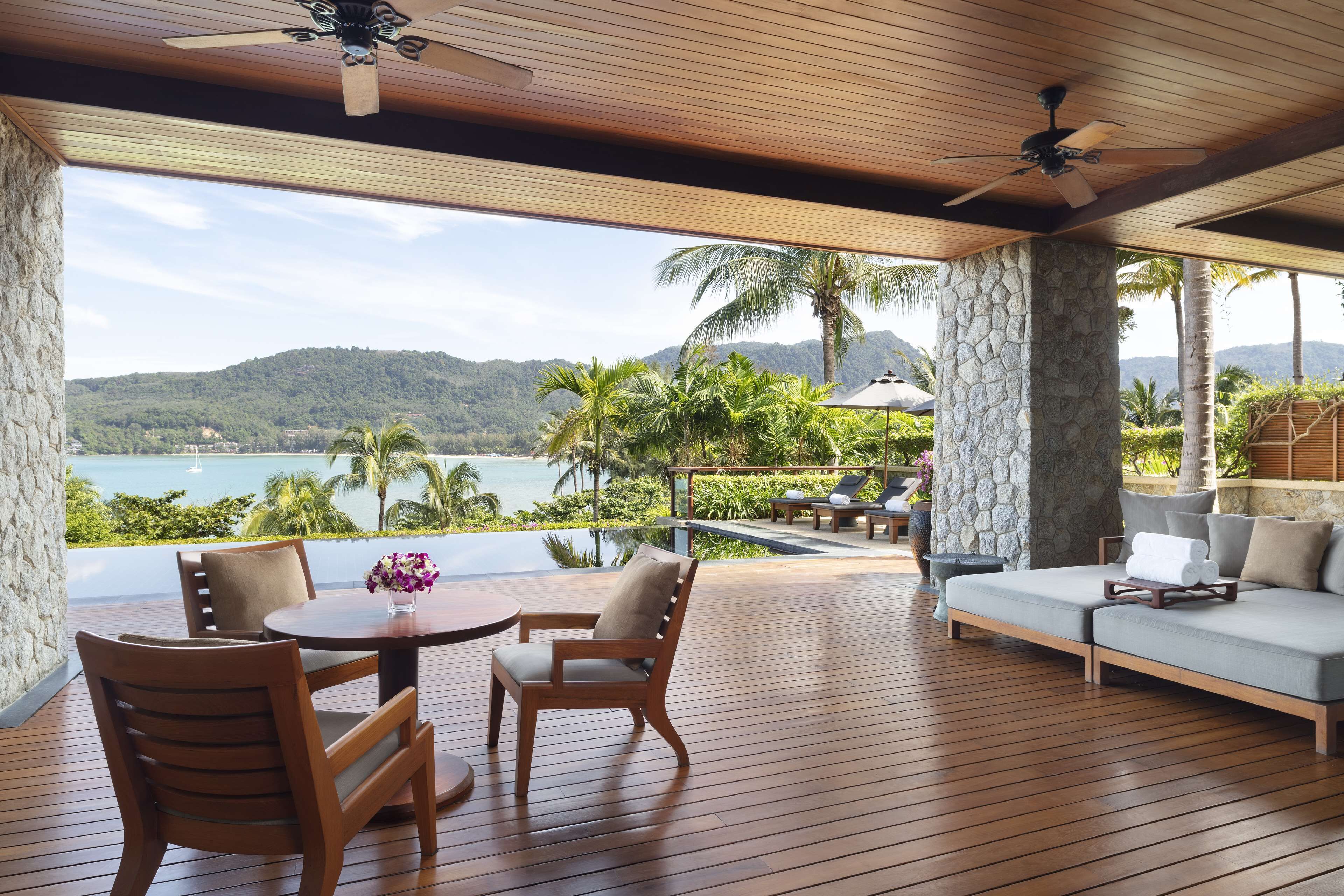 A wooden deck with a table and chairs, a sofa, and a view of a body of water and mountains.