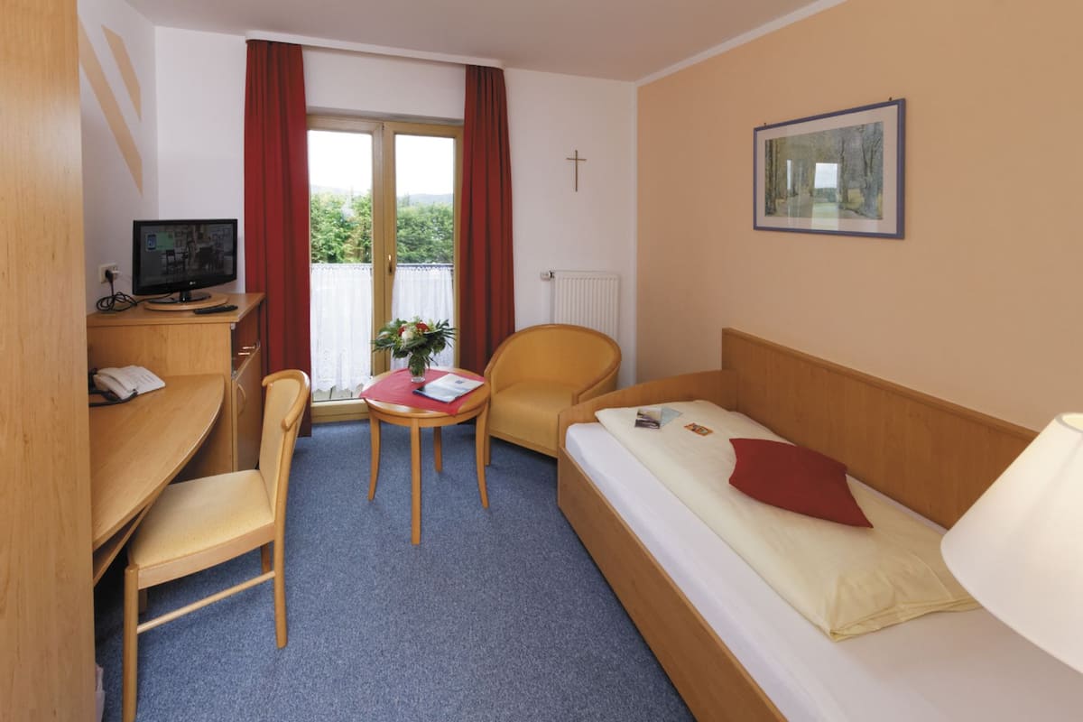 Habitación de hotel con cama, escritorio con computadora, silla, pequeña mesa con un florero y ventana con cortinas.