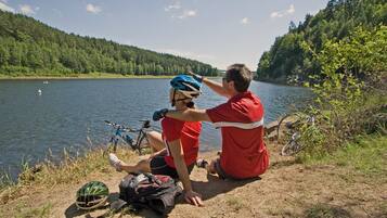 A person sitting by a lake with a bicycle and a helmet, another person adjusting the helmet, and a backpack on the ground.