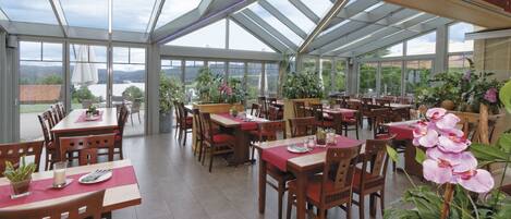 A spacious indoor dining area with multiple tables and chairs, large windows, and potted plants.
