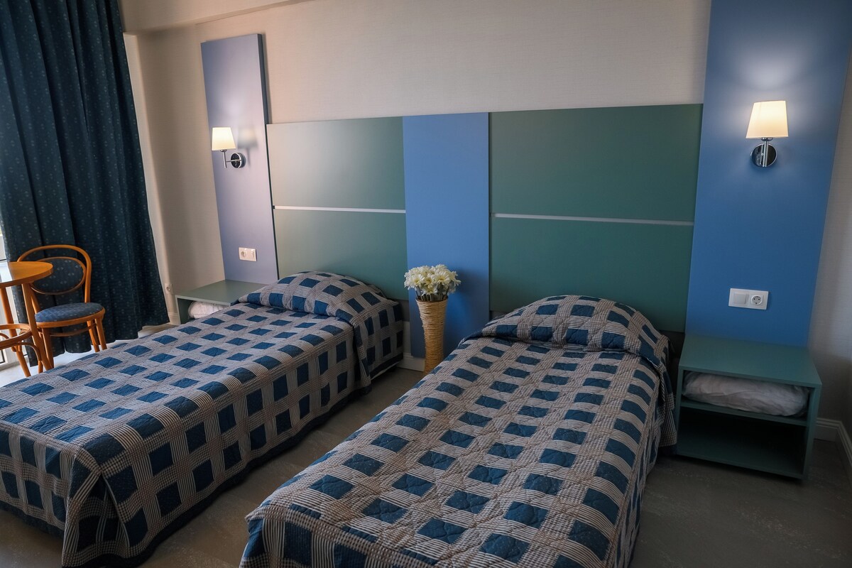 A hotel room with two single beds, a small table, a chair, a vase with flowers, and a wall-mounted lamp.