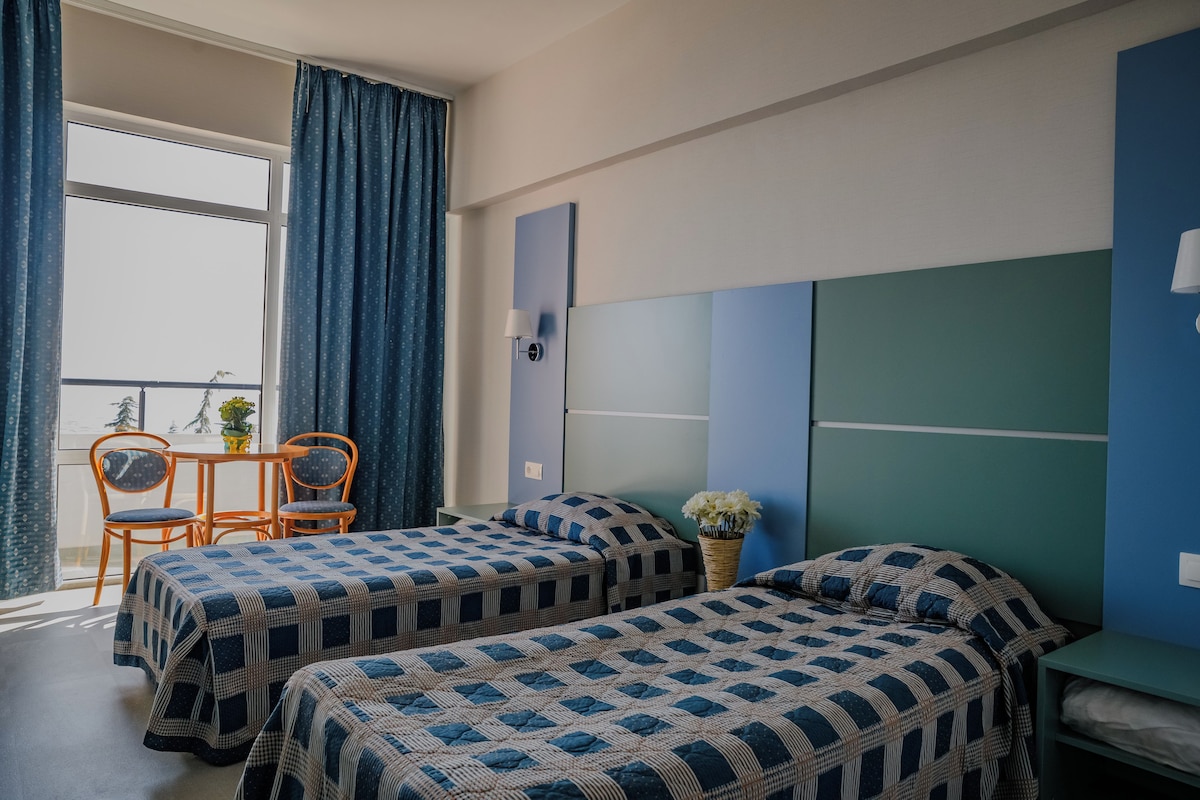Une chambre avec deux lits, une table avec des chaises, une fenêtre avec des rideaux et une lampe fixée au mur.