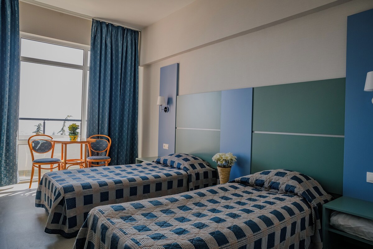 A room with two beds, a table with chairs, a window with curtains, and a wall-mounted lamp.