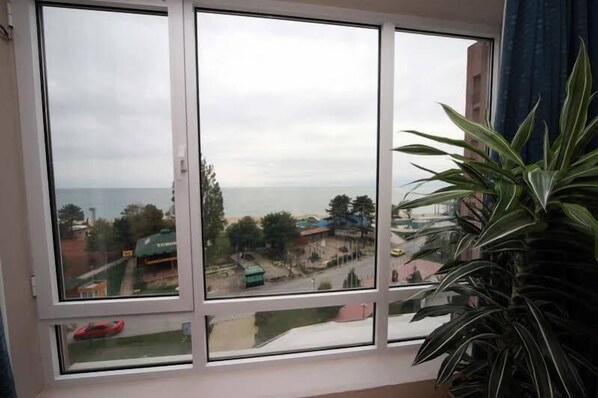 A room with a large window overlooking a coastal area, a potted plant, and a red car in the view. - Hotel Lilia (Varna)