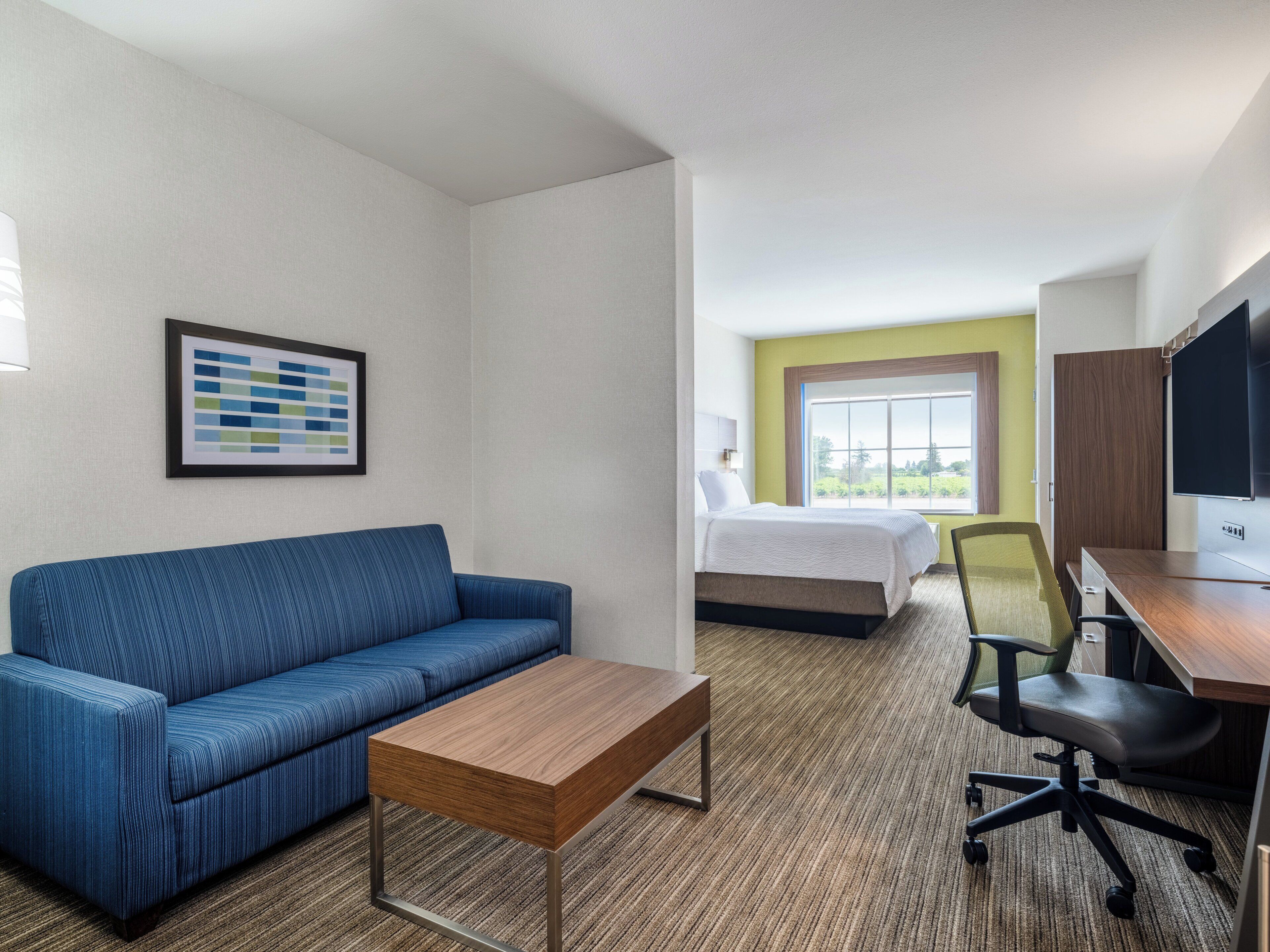 A hotel room with a blue sofa, a wooden coffee table, a bed with white bedding, a desk with a chair, and a television.