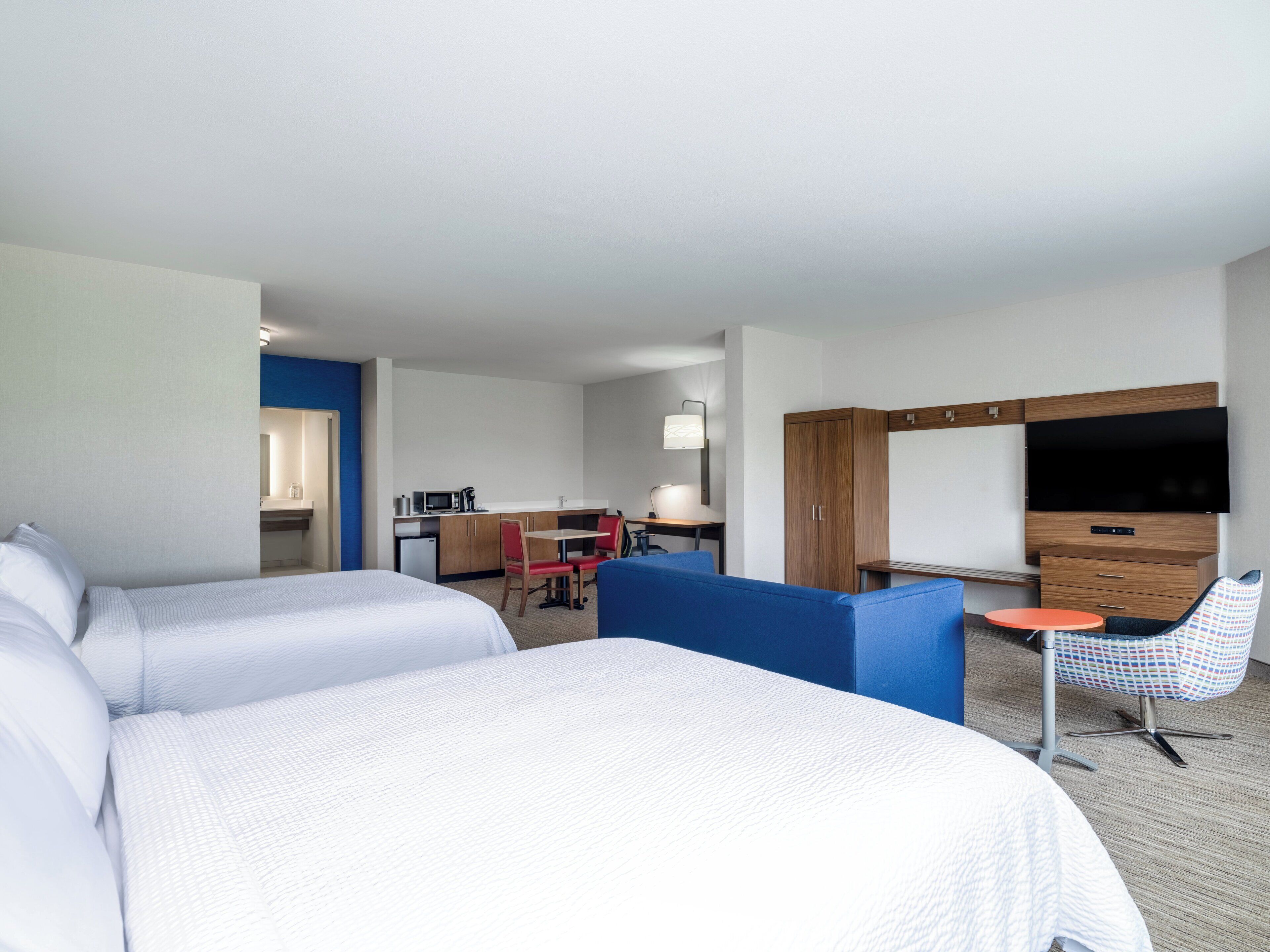 A hotel room with two beds, a kitchenette, a TV, and a seating area with a chair and table.