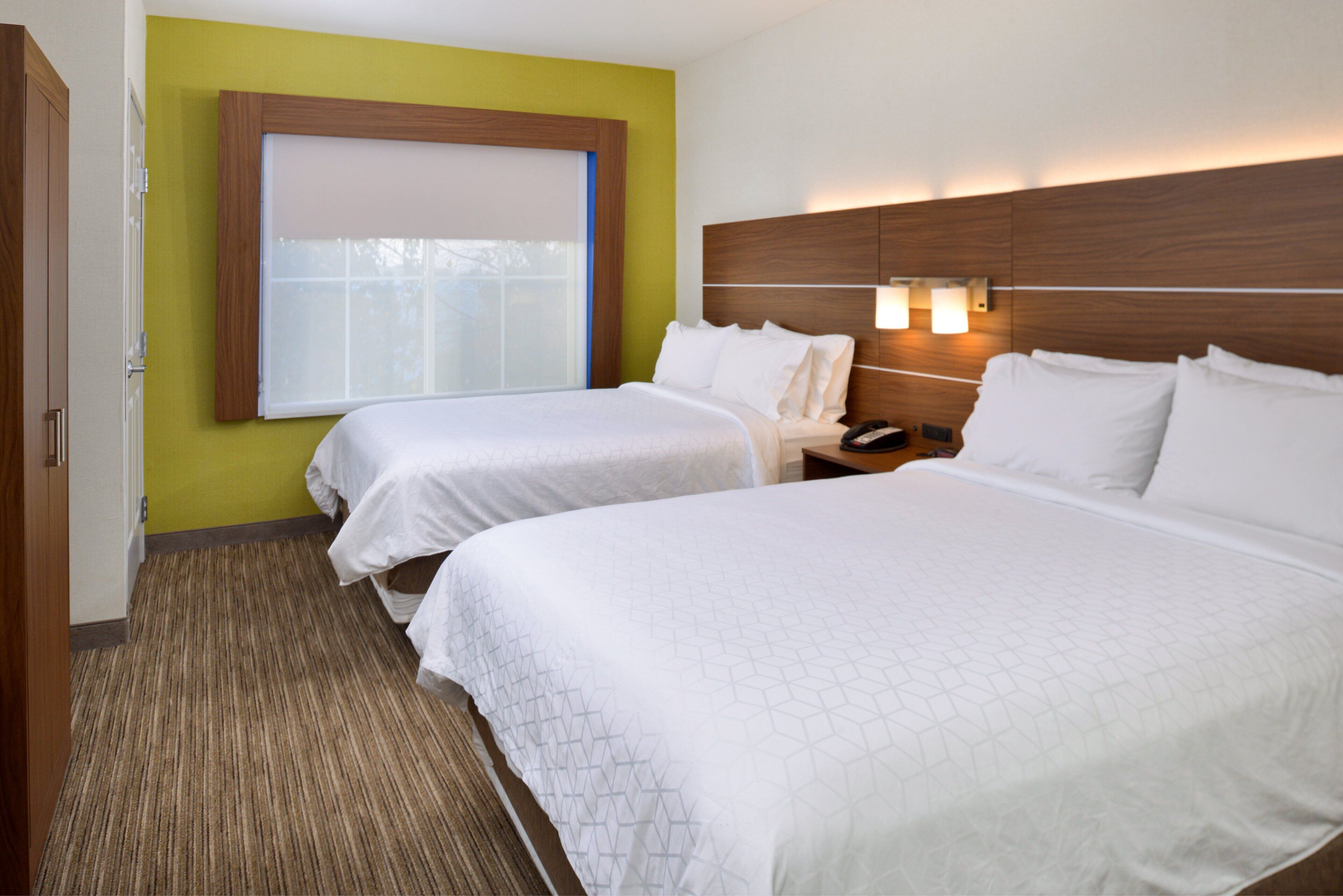 A hotel room with two beds, a window with blinds, a nightstand with a phone, and a wall-mounted light fixture.