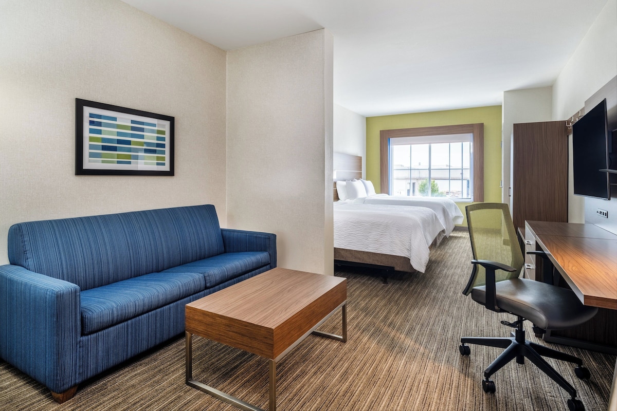 A hotel room with a blue sofa, a wooden coffee table, a bed with white bedding, a desk with a chair, and a TV on the wall.