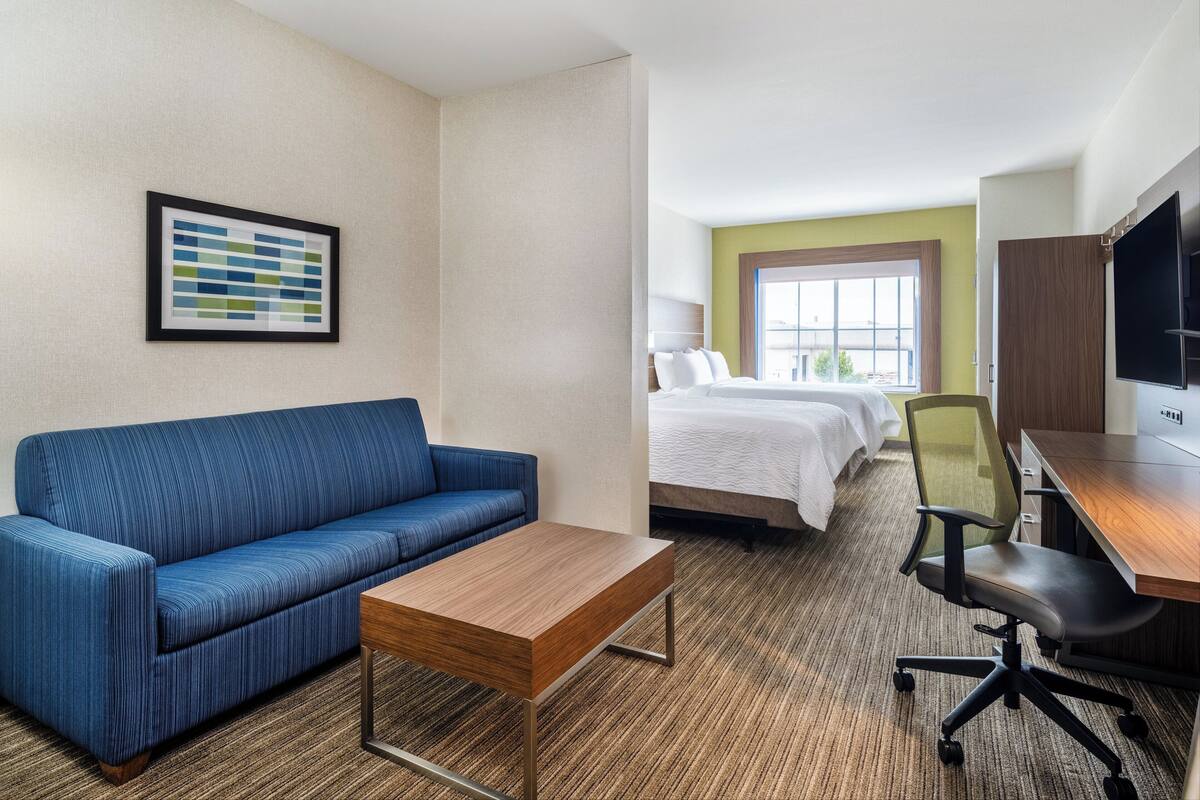 A hotel room with a blue sofa, a wooden coffee table, a bed with white bedding, a desk with a chair, and a TV on the wall.