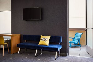 A modern room with a dark blue bench, a yellow pillow with "POW!" text, a wooden desk, a yellow chair, and a blue chair. - Aloft by Marriott Leawood-Overland Park (Leawood)