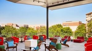 Un patio al aire libre con diferentes opciones de asientos, incluyendo sillas de madera y sofás rojos, una sección cubierta con mesas, y vistas de edificios y árboles circundantes.