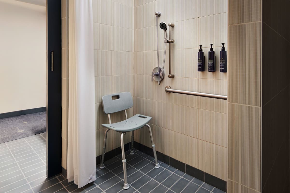 A bathroom with a shower, a grab bar, a wheelchair-accessible chair, and toiletries on the wall.