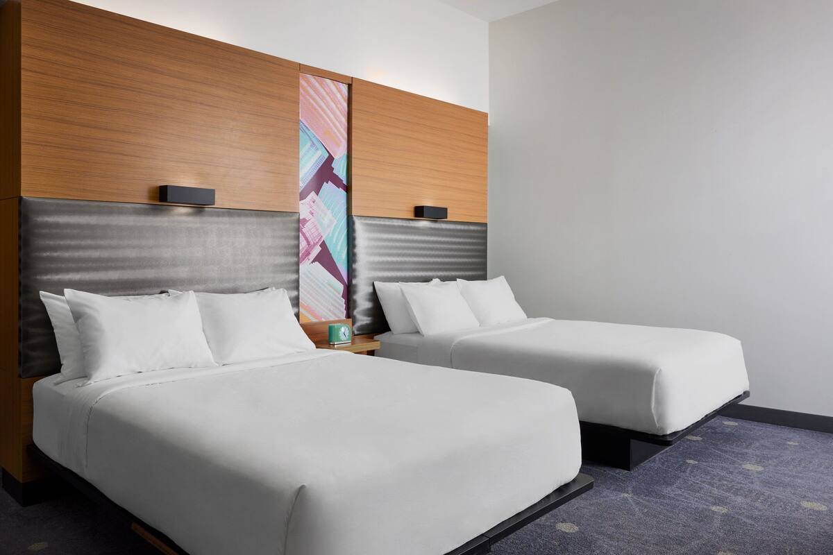 Ein Hotelzimmer mit zwei Betten, einem Holz-Kopfteil, einem farbenfrohen abstrakten Kunstwerk und einer kleinen Uhr auf dem Nachttisch.