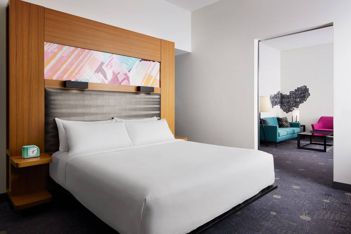 Ein Hotelzimmer mit einem großen Bett, einem Holzkopfstück, einem Nachttisch, einer Uhr und einem farbenfrohen, abstrakten Kunstwerk über dem Bett.