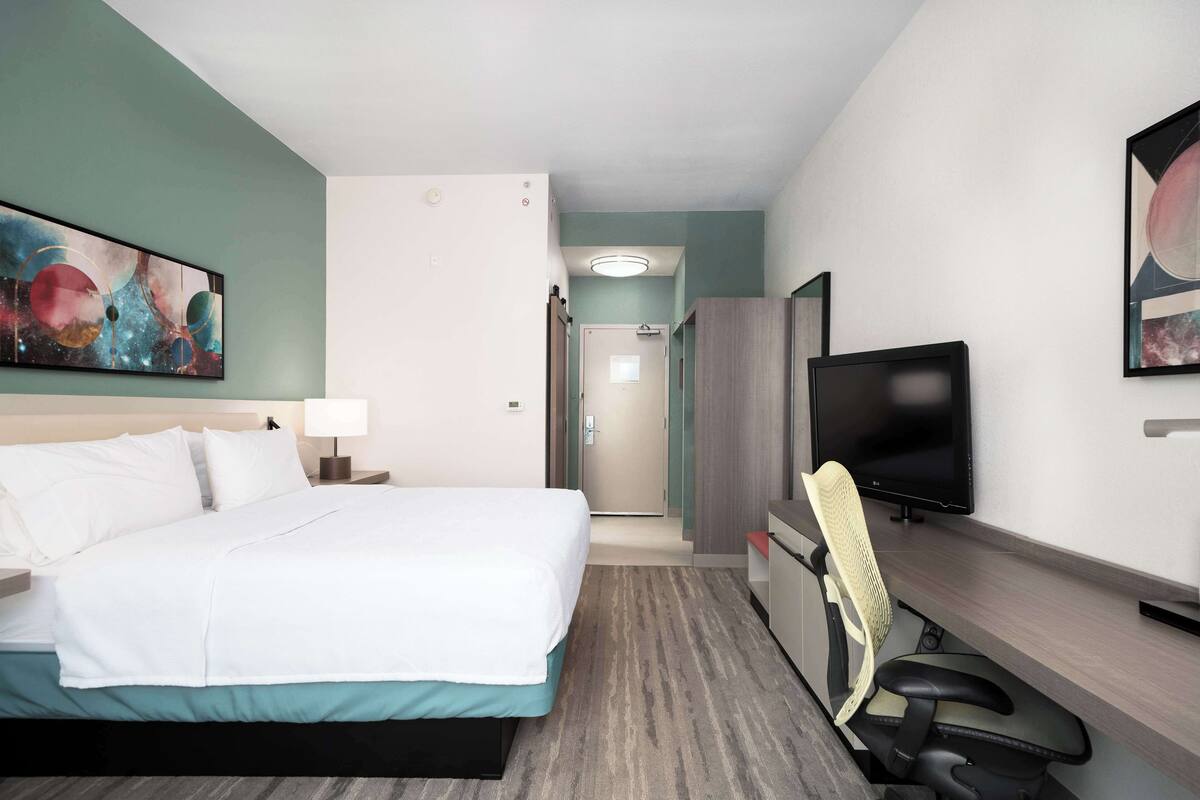 A hotel room with a bed, a desk with a chair, a television, and artwork on the walls.